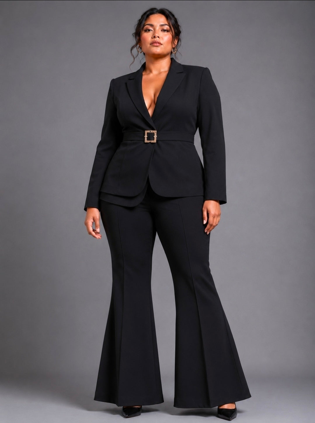 A woman modeling a black two-piece suit with a belted waist and flared pants, standing against a solid gray studio background.