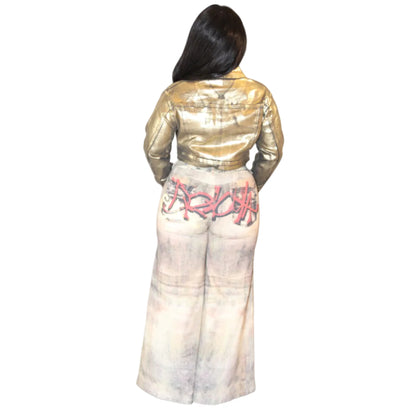 Woman wearing a gold jacket and light colored jeans on a white background. The jeans are detailed with multicolored graffiti. (back)