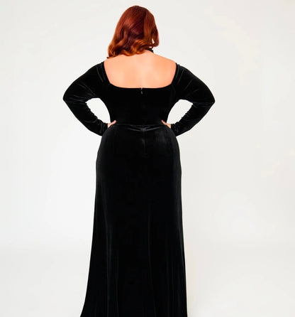 A woman with red hair is shown from the back wearing a long black velvet gown with long sleeves and a square neckline, standing against a plain light gray studio background.