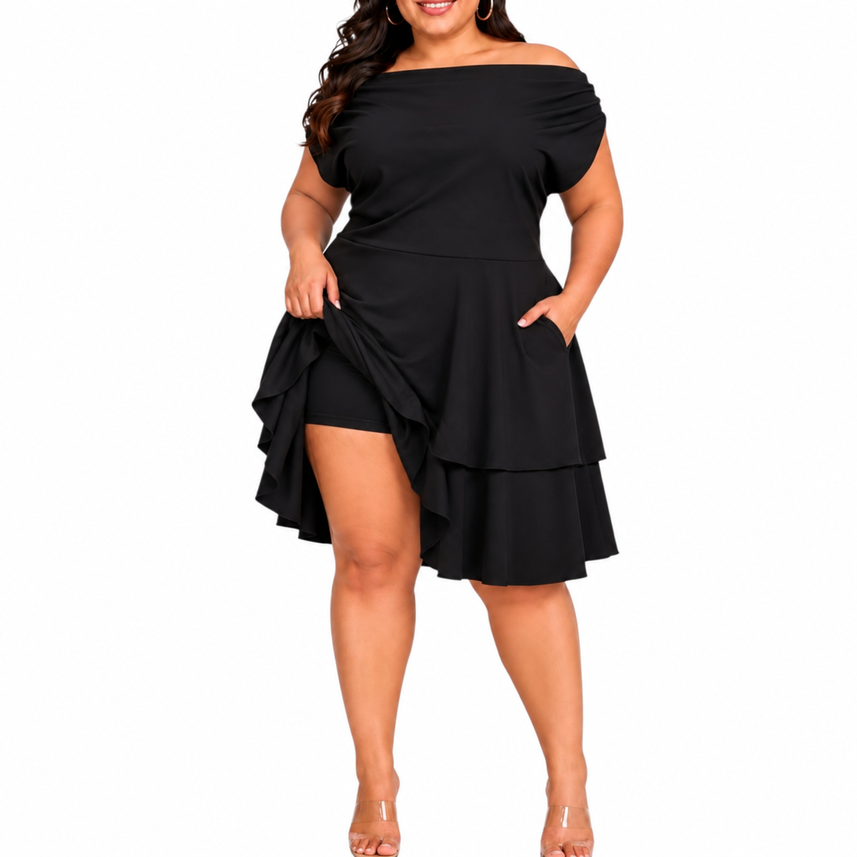 A woman modeling a black off-the-shoulder  dress with a layered ruffle skirt and attached shorts, paired with clear high-heeled sandals, against a white studio background