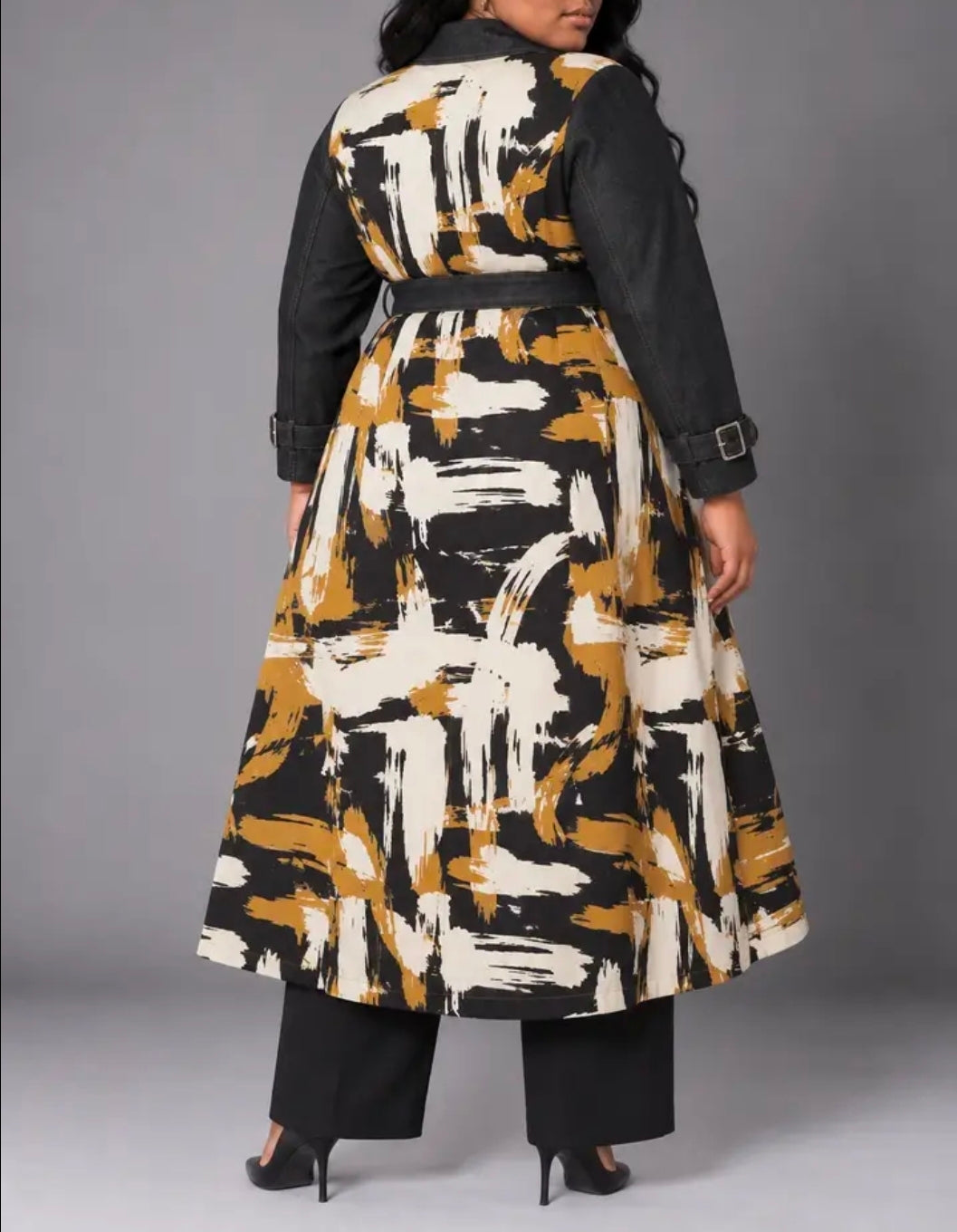 A woman modeling a long black trench-style coat with a bold abstract brushstroke pattern in black, white, and mustard yellow. The coat features long black sleeves, a matching black belt, and is shown from the back against a solid gray studio background.