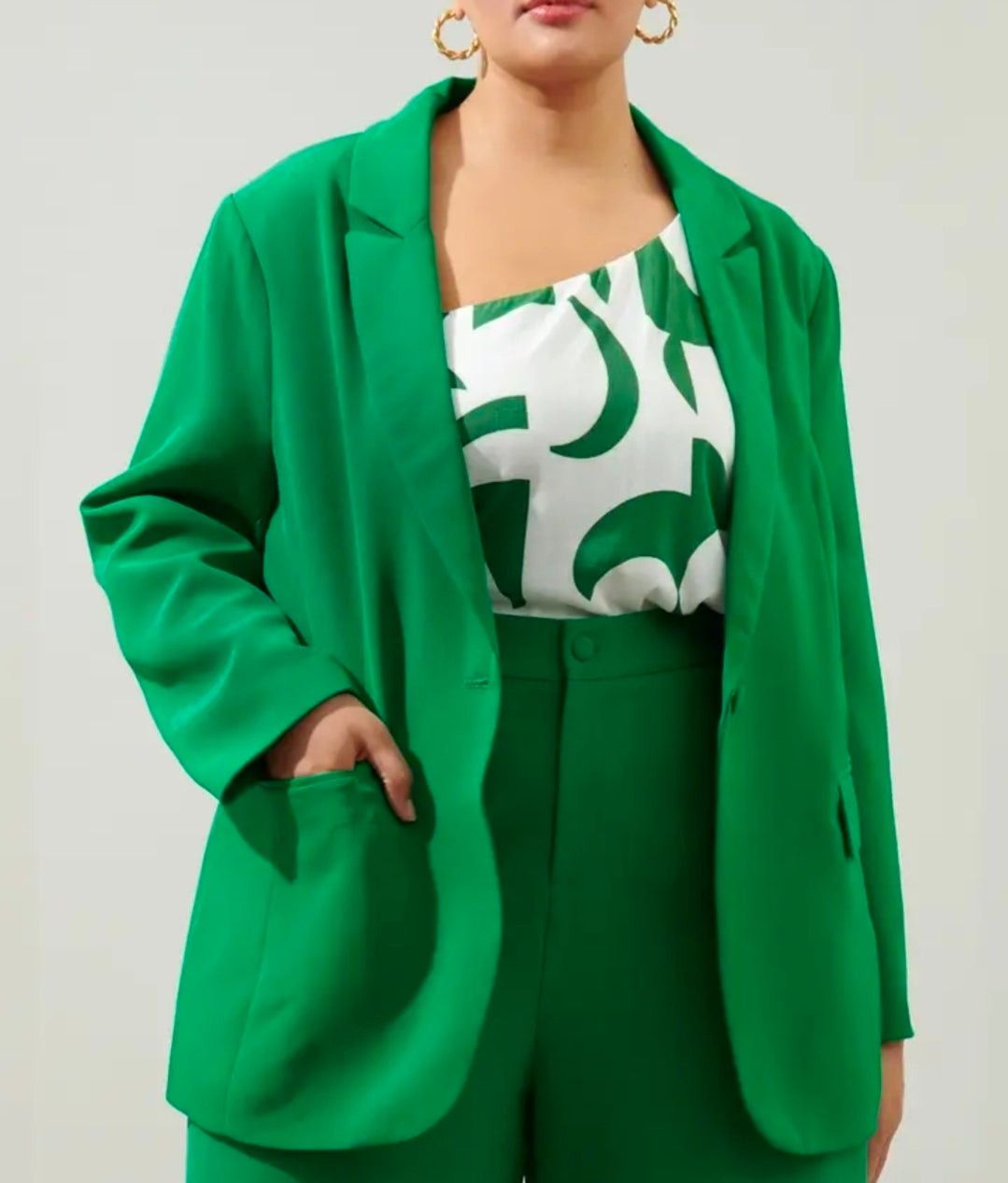 A model wearing a bright green tailored blazer over a white top with a green abstract pattern, paired with matching green trousers, against a light gray studio background.
