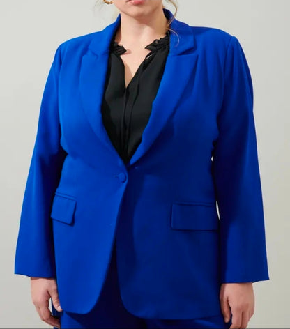 A woman wearing a bright royal blue tailored blazer over a black blouse, shown from the lower face to the hips against a plain light gray studio background.