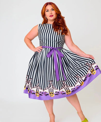 A woman modeling a sleeveless A-line dress with black and white vertical stripes, a purple waist tie belt, and a purple hem featuring a pumpkin and skull pattern, set against a plain light gray background.