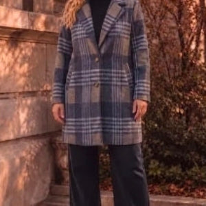 A person wearing a long plaid coat in navy, gray, and beige tones, standing outdoors against a stone wall and trees.