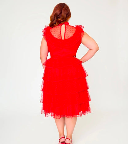 A woman stands with her back to the camera wearing a bright red tiered tulle dress with a sheer polka-dot back panel and ruffled cap sleeves, paired with pink sandals, against a plain white studio background.