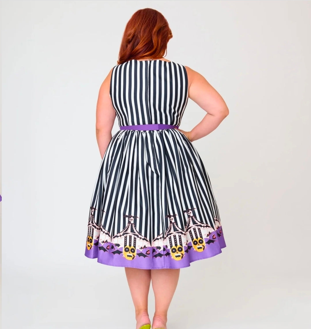 A woman viewed from the back wearing a sleeveless black and white vertically striped dress with a purple waistband and a purple hem featuring a Halloween-themed print of skeletons, bats, and pumpkins.