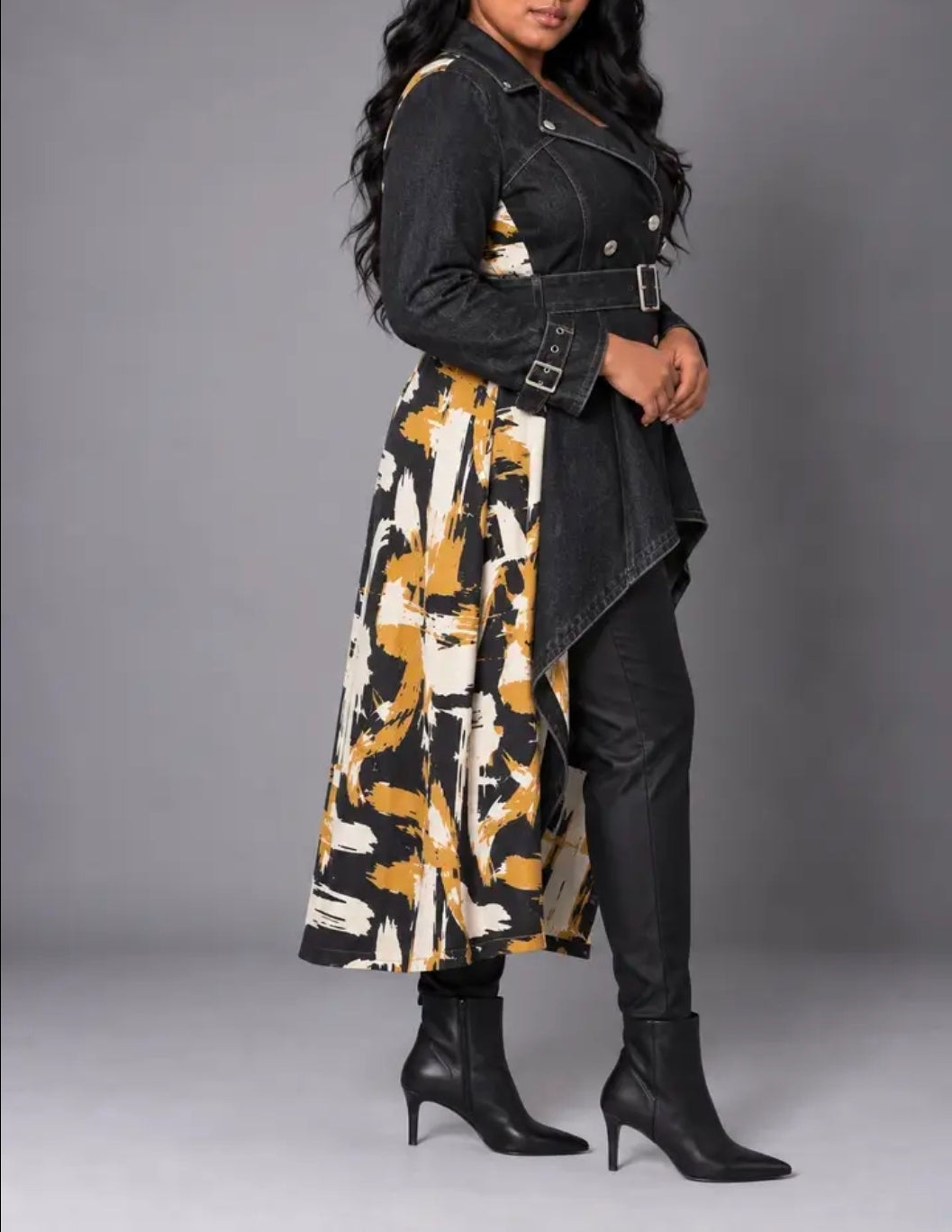A woman models a long black denim-style jacket with a belted waist, worn over a black, white, and mustard yellow abstract print dress, paired with black pointed-toe heeled ankle boots, against a solid gray studio background.